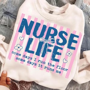 May include: White sweatshirt with a graphic that reads "NURSE LIFE" in blue, with pink vertical stripes. The text "some days I run the floor some days it runs me" is below the main text. Small heart and flower graphics are also included.