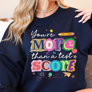 May include: Navy blue sweatshirt with the text "You're More than a test SCORE" in colorful, playful lettering. The design includes stars, a pencil, and other school-themed graphics.