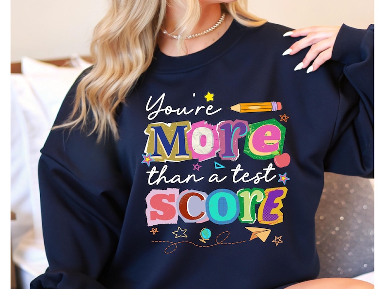 May include: Navy blue sweatshirt with the text "You're More than a test SCORE" in colorful, playful lettering. The design includes stars, a pencil, and other school-themed graphics.
