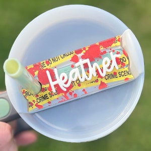 May include: A white plastic cup lid with a red and yellow crime scene tape sticker that says "Heather" on it. The sticker has red blood splatter and a green background.