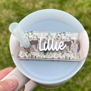 May include: A white and pink travel mug with a straw and a personalized name tag. The tag has a floral pattern with a pink background and white goats. The name "Lillie" is written in white on the tag.