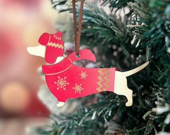 May include: A wooden Christmas ornament shaped like a dachshund dog. The dog is wearing a red sweater with snowflake designs and a gold zig-zag pattern. The ornament hangs from a brown string and is displayed on a Christmas tree.