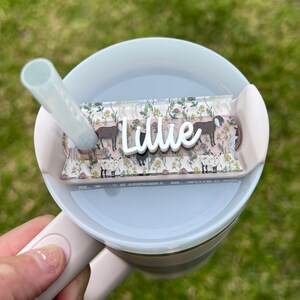 May include: A pink and white tumbler with a clear lid and a straw. The lid has a clear plastic insert with a floral pattern and the name "Lillie" written in white.