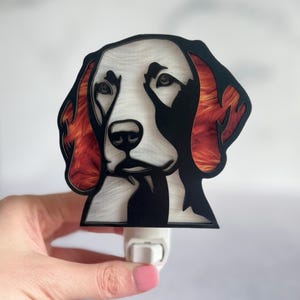 May include: A decorative night light featuring a dog's face. The dog's face is white and black, with reddish-orange accents on the ears. The night light is plugged into a white outlet.