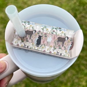 May include: A pink and white tumbler with a clear lid and a straw. The lid has a removable insert with a floral pattern featuring sheep and goats.