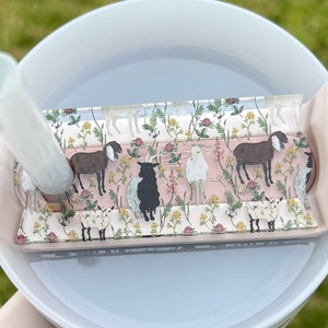 May include: A white ceramic mug with a pink silicone lid and a clear plastic insert featuring a floral pattern with goats. The insert has a pink background with green leaves and flowers. The goats are brown, white, and black.