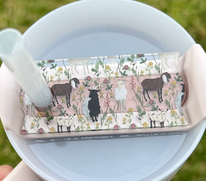 May include: A white ceramic mug with a pink silicone lid and a clear plastic insert featuring a floral pattern with goats. The insert has a pink background with green leaves and flowers. The goats are brown, white, and black.