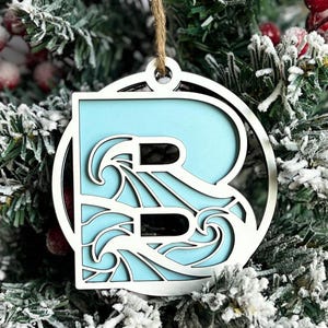 May include: A light blue wooden ornament shaped like the letter B with a wave design cut out. The ornament is hanging on a Christmas tree.