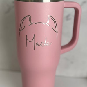 Puede incluir: Una taza de viaje de acero inoxidable rosa con tapa negra y asa. La taza tiene un dibujo lineal de un perro con el nombre "Mack" grabado en ella.