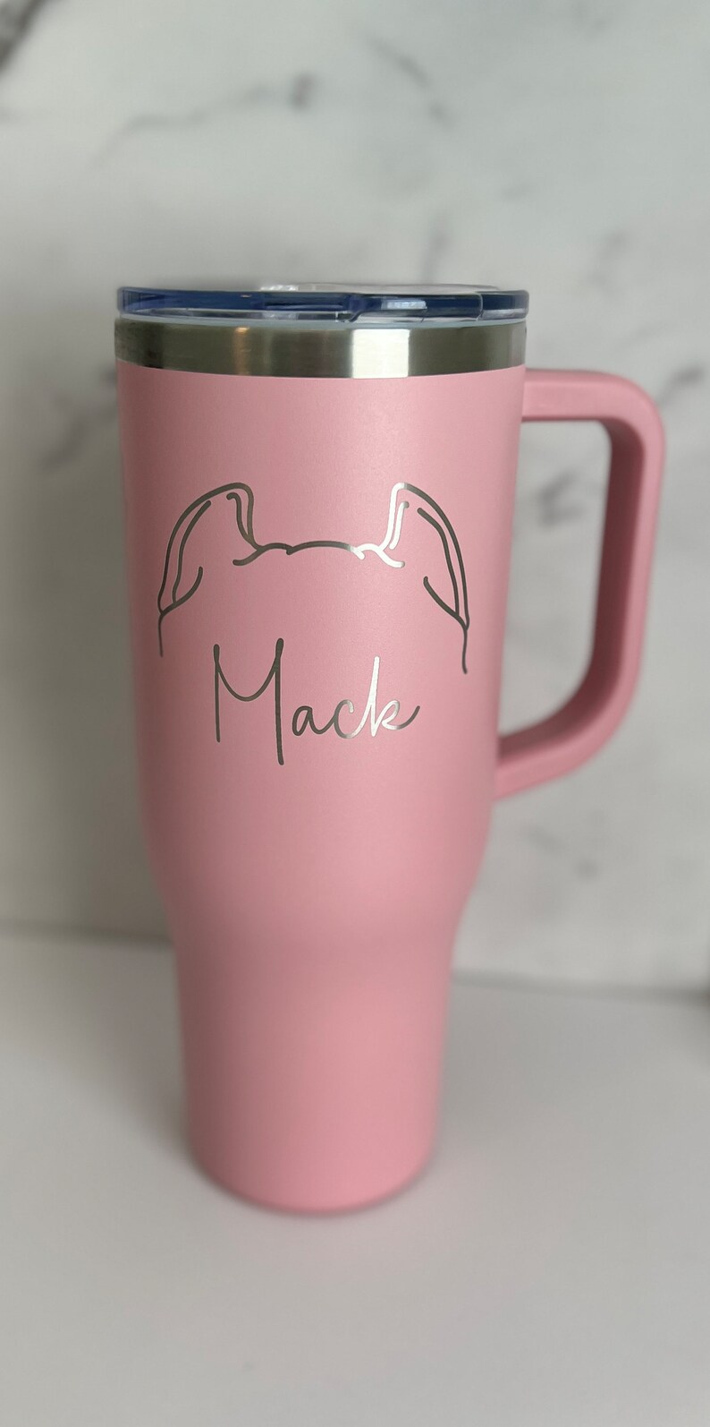 Puede incluir: Una taza de viaje de acero inoxidable rosa con tapa negra y asa. La taza tiene un dibujo lineal de un perro con el nombre "Mack" grabado en ella.