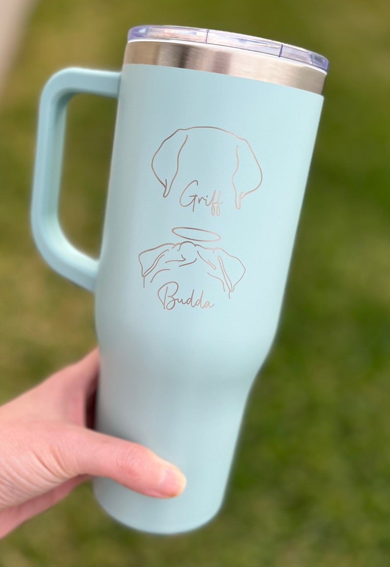 Puede incluir: Una taza de viaje de acero inoxidable azul claro con un asa negra. La taza tiene un contorno blanco de un perro con el nombre "Griff" y un contorno blanco de un carlino con un halo y el nombre "Buddha" debajo.