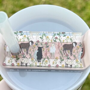 May include: A close-up of a clear plastic lid with a pink and green floral design featuring goats. The lid is on top of a white tumbler with a straw.