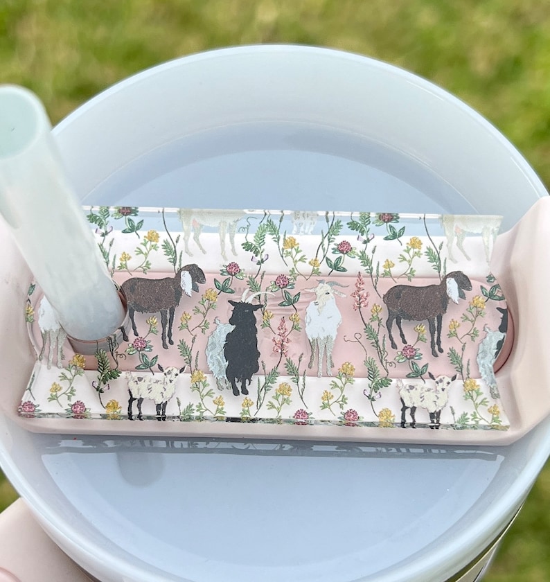 May include: A close-up of a clear plastic lid with a pink and green floral design featuring goats. The lid is on top of a white tumbler with a straw.