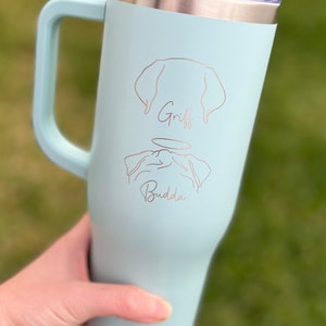 Puede incluir: Una taza de viaje de acero inoxidable azul claro con asa. La taza tiene un dise&ntilde;o grabado con l&aacute;ser de un perro con los nombres "Griff" y "Buddha" debajo.