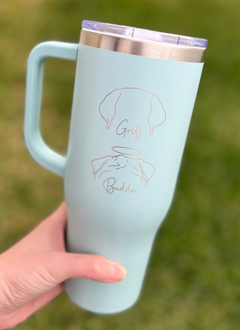 Puede incluir: Una taza de viaje de acero inoxidable azul claro con asa. La taza tiene un dise&ntilde;o grabado con l&aacute;ser de un perro con los nombres "Griff" y "Buddha" debajo.