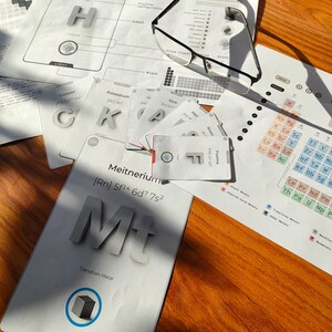 Pode incluir: Uma coleção de materiais educativos, incluindo cartões e diagramas da tabela periódica, está disposta sobre uma superfície de madeira. Os cartões apresentam símbolos de elementos como 'Mt' para Meitnerium e 'K' para Potássio. Um par de óculos repousa sobre os papéis.