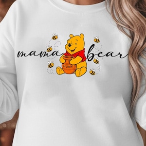 May include: White sweatshirt featuring a graphic of Winnie the Pooh holding a honey pot, surrounded by bees. The words "mama bear" are written in cursive. The bear is wearing a red shirt.