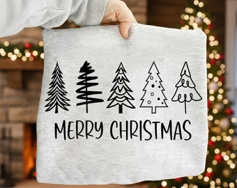 Könnte beinhalten: Ein hellgraues Sweatshirt mit fünf schwarzen Weihnachtsbäumen und dem Schriftzug "MERRY CHRISTMAS". Das Sweatshirt wird von einer Hand gehalten, mit einem Kamin und einem Weihnachtsbaum im Hintergrund.