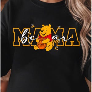 May include: Black sweatshirt featuring a graphic of Winnie the Pooh holding a honey pot. The text "Mama Bear" is displayed in a combination of block and script fonts, with bees scattered around the text and character.
