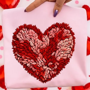 May include: A light pink fabric with a large, textured heart design in shades of red and pink. The heart appears to be made of yarn or a similar material. Red heart-shaped balloons are in the background.