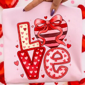 Könnte beinhalten: Ein rosa Sweatshirt mit dem Wort "LOVE" in roten und weißen Buchstaben. Das "L" ist mit kleinen roten Herzen gefüllt, das "O" ist ein gestreiftes Herz mit einer roten Schleife, das "V" ist mit roten Herzen gefüllt und das "E" ist mit roten Herzen gefüllt. Kleine rote Herzen sind verstreut.