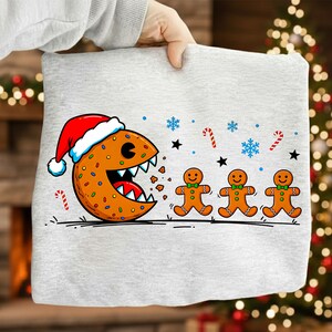 Può includere: Felpa grigio chiaro con un design a cartoni animati festivo. Raffigura un biscotto di pan di zenzero con un cappello di Babbo Natale che mangia omini di pan di zenzero, con fiocchi di neve, stelle e bastoncini di zucchero. Un design divertente e stagionale.