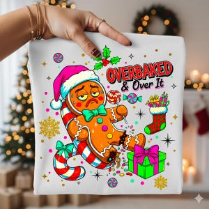 May include: White Christmas-themed graphic featuring a dejected gingerbread man wearing a pink Santa hat and holding a lollipop. The text "OVERBAKED & Over It" is in red. The design includes a candy cane, gift, and stocking.