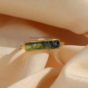 Puede incluir: Un anillo de oro con una piedra rectangular verde y negra. La piedra está engastada en una banda de oro, creando un diseño moderno. El anillo se exhibe sobre una tela color melocotón. El diseño del anillo es elegante y contemporáneo.