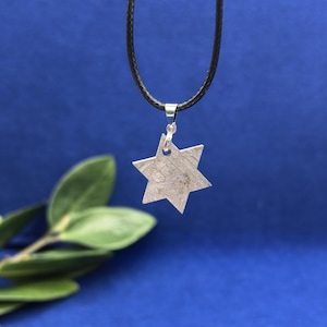 Puede incluir: Un colgante plateado en forma de estrella de seis puntas cuelga de un collar de cordón negro. La estrella tiene una superficie texturizada y está suspendida de una pequeña anilla plateada. El fondo es azul oscuro, con hojas verdes.