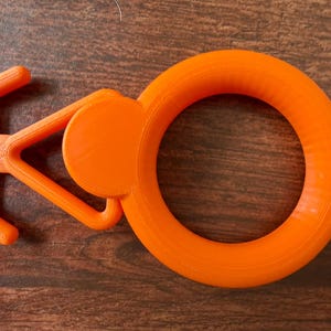 May include: A vibrant orange 3D-printed item featuring a circular ring and a handle-like design. The handle has a triangular form with a rounded disc attached. The object is made of plastic and rests on a brown wooden surface.