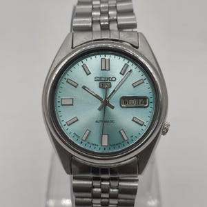 May include: A silver-toned Seiko 5 automatic wristwatch with a light blue dial. The watch features silver hands and markers, a day-date display, and a stainless steel band. The watch face reads "Seiko 5", "Automatic", and "Made in Japan".