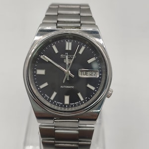 Pode incluir: Um relógio de pulso automático Seiko prateado com mostrador cinza escuro. O relógio apresenta um visor de dia e data, ponteiros e marcadores luminosos e uma pulseira de aço inoxidável. O mostrador do relógio tem aproximadamente 3,8 cm de diâmetro.