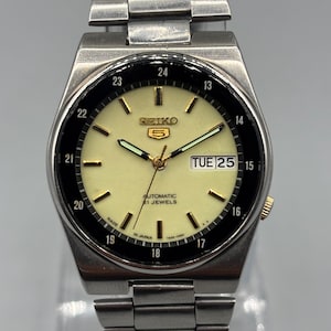May include: A silver-toned Seiko automatic wristwatch with a cream-colored face and a black bezel. The watch displays the day and date, with gold-toned hands and markers. The watch band is made of silver-toned metal links.