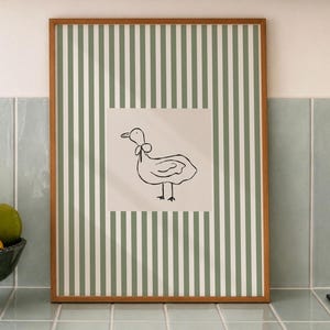 May include: A rectangular wall art print in a wooden frame featuring a vertical sage green and white striped pattern with a central white square containing a simple black line drawing of a duck wearing a small bow around its neck.