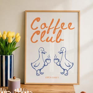 Könnte beinhalten: Ein gerahmter Kunstdruck mit dem orangefarbenen Schriftzug Coffee Club über zwei blauen Enten-Strichzeichnungen, die Kaffeetassen halten, mit dem Text OPEN DAILY darunter. Der Druck steht auf einem Holztisch mit gestreiften Tassen und einem Croissant.