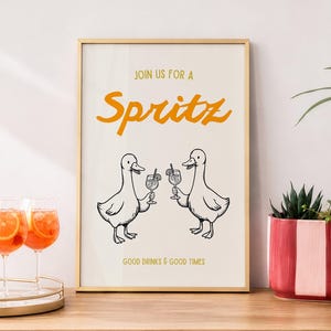 Könnte beinhalten: Ein gerahmter Kunstdruck mit cremefarbenem Hintergrund und dem orangefarbenen Text JOIN US FOR A SPRITZ. Darunter halten zwei schwarz-weiße Enten-Strichzeichnungen Cocktailgläser. Der untere Text lautet GOOD DRINKS & GOOD TIMES in Orange.