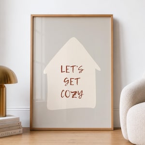 Könnte beinhalten: Ein rechteckiges Wandbild in einem hellen Holzrahmen mit einer beigen, hausförmigen Silhouette auf hellgrauem Hintergrund. Der Text "LET'S GET COZY" ist in einer dunkelbraunen, handschriftlichen Schriftart in der Hausform gedruckt.