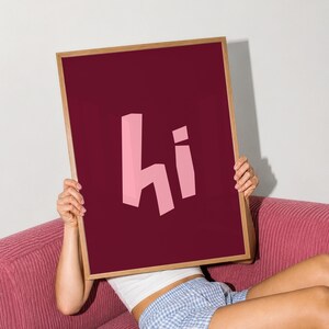 Könnte beinhalten: Ein rechteckiges gerahmtes Wandbild mit einem tief burgunderroten Hintergrund und dem Wort hi in einer stilisierten, blockartigen hellrosa Schrift. Der Rahmen besteht aus hellem Holz und hält den Druck sicher in seinen dünnen, sauberen Kanten.