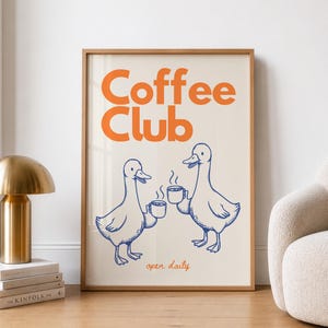 Könnte beinhalten: Ein gerahmter Kunstdruck mit den Worten "Coffee Club" in Orange, über zwei blau gezeichneten Enten, die Kaffeetassen halten. Die Worte "open daily" stehen darunter. Der Druck ist in einem hellen Holzrahmen.
