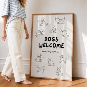 Könnte beinhalten: Ein gerahmter Druck mit den Worten "DOGS WELCOME" und dem Satz "humans may enter too." Der Druck zeigt schwarz-weiße Strichzeichnungen verschiedener Hunderassen. Der Rahmen hat eine hellbraune Farbe.