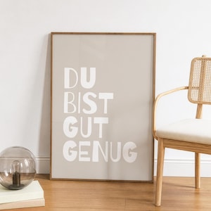 Könnte beinhalten: Ein rechteckiges Wandbild in einem dünnen Holzrahmen mit hellbeigem Hintergrund und dem weißen Schriftzug "DU BIST GUT GENUG" in einer fetten, abgerundeten serifenlosen Schrift. Das Bild ist im Hochformat ausgerichtet.