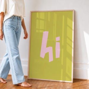 Könnte beinhalten: Ein gerahmter Kunstdruck mit einem hellen Holzrahmen. Das Kunstwerk zeigt das Wort "hi" in Rosa auf einem hellgrünen Hintergrund. Der Druck lehnt an einer weißen Wand.