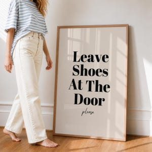 Könnte beinhalten: Ein gerahmter Druck mit dem Text "Leave Shoes At The Door please" in schwarzer Schrift. Der Druck hat einen beigefarbenen Hintergrund und einen Holzrahmen. Eine Person mit gestreiftem Hemd und cremefarbener Hose steht neben dem Druck.