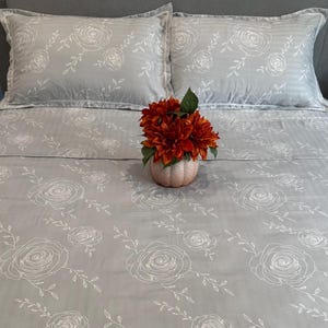 May include: A gray bed set with a white rose pattern, including two pillowcases and a fitted sheet. A small orange floral arrangement in a pink pumpkin-shaped vase sits on the bed. The bedding has a striped texture.