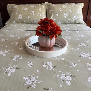May include: A round white tray with clear handles and a mirrored base sits on a sage green bedspread featuring a white floral pattern. A small, textured, light-colored vase holds a bouquet of deep orange flowers, centered on the mirrored tray.
