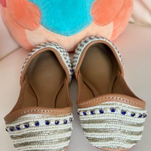 May include: A pair of white beaded flats with brown leather insoles. The flats have a striped pattern with silver and blue beads. A plush orange cat-shaped pillow with a blue heart is in the background.