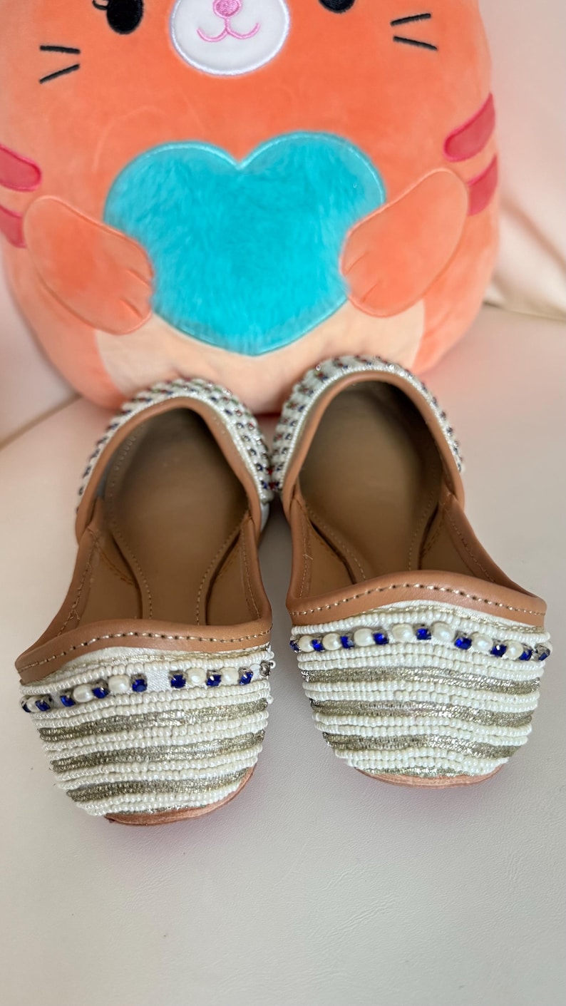 May include: A pair of white beaded flats with brown leather insoles. The flats have a striped pattern with silver and blue beads. A plush orange cat-shaped pillow with a blue heart is in the background.