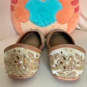 May include: A pair of cream-colored slip-on shoes with intricate gold embroidery and embellishments. The shoes have a tan leather trim and a small heel. A plush orange cat-shaped pillow with a blue heart is in the background.