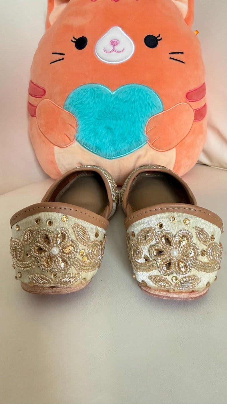 May include: A pair of cream-colored slip-on shoes with intricate gold embroidery and embellishments. The shoes have a tan leather trim and a small heel. A plush orange cat-shaped pillow with a blue heart is in the background.