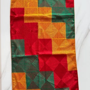 May include: A rectangular table runner featuring a geometric pattern of squares in vibrant red, emerald green, and golden yellow. The fabric displays a textured, stitched grid design across the entire surface, creating a repeating block motif.